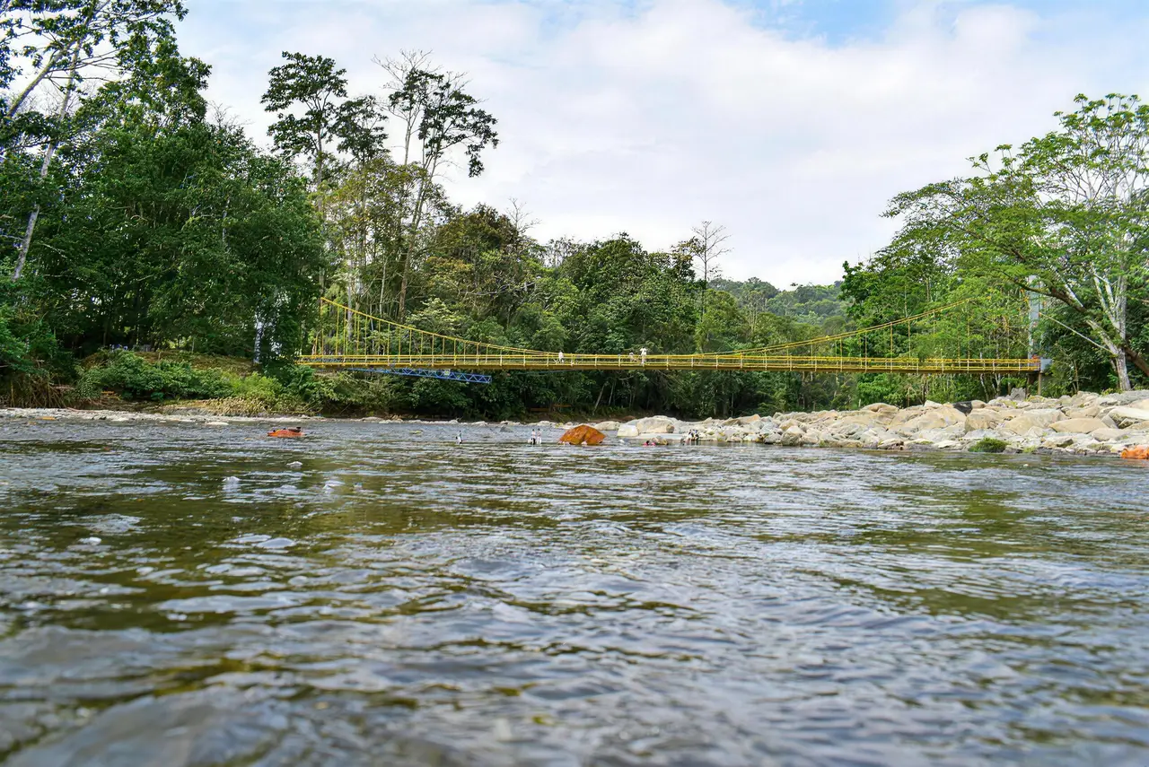 Río de aguas claras entre piedras en la vereda Caracolí, zona rural de Apartadó, con un puente colgante amarillo al fondo.